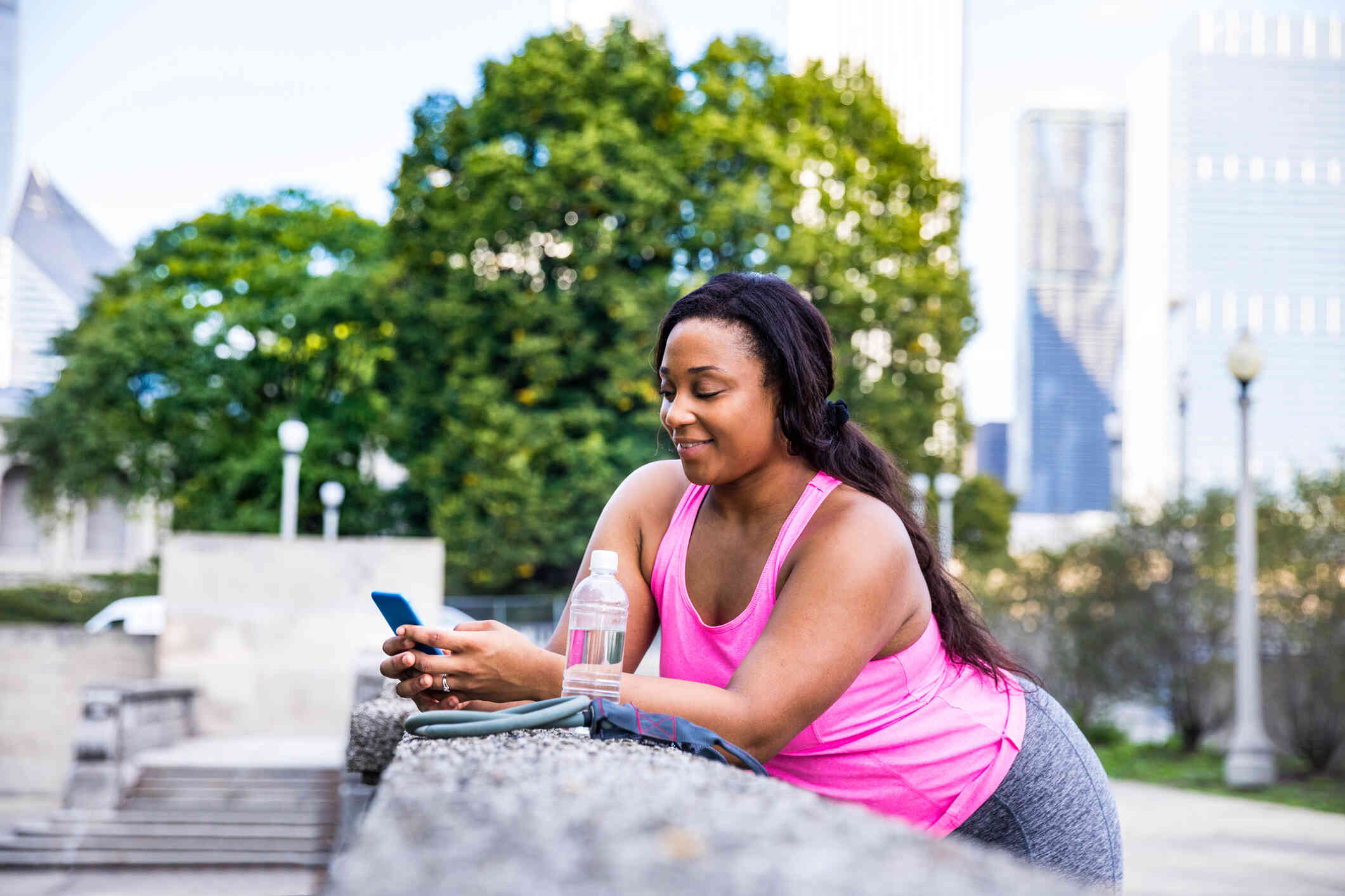 A woman in a pink workout shirt leans against a railing with a bottle of water whole holding her phone.